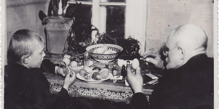 Fotonuotrauka. Grupinė. Senelis (dešinėje) su anūku (kairėje) dažo velykinius margučius prie uždaroje patalpoje esančio stalo. XX a. 3–4 deš. (Trakų muziejus, CC BY)